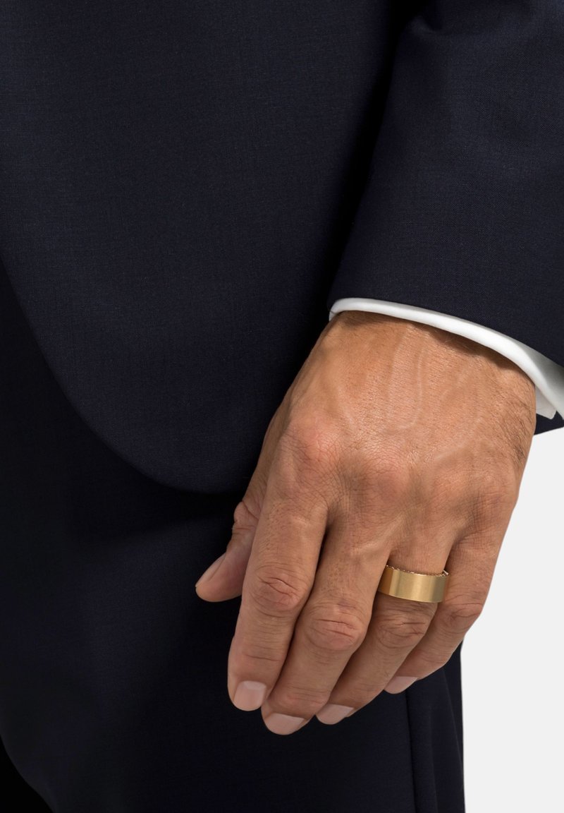 Goldener Metalring an einer Hand mit heller Haut, die einen dunklen Anzug mit einem weiß geknoteten Hemd, das am Bund sichtbar ist, trägt. Glatte Textur, rechteckige Form.