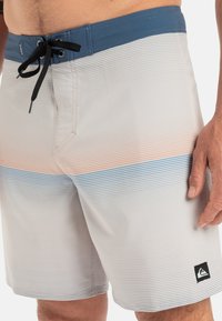 Herrliche Boardshorts in Grau mit horizontalen Streifen in Orange und Blau. Sie verfügen über einen blauen Bund, einen schwarzen Kordelzug und ein seitliches Logo-Patch.