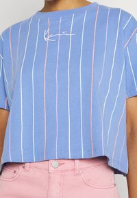 Beskuren blå t-shirt med vertikala ränder i rosa, vitt och lavendel. Har en rundad halsringning och en broderad logotyp på bröstet.