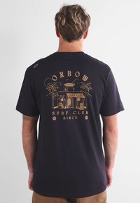 T-shirt en coton noir avec un motif rétro de club de surf doré, comprenant des palmiers et du texte. Manches courtes, coupe décontractée, texture douce.
