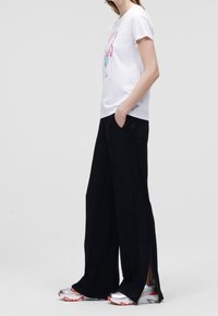 Camiseta blanca con gráfico colorido, pantalones anchos negros con patrón texturizado y zapatillas deportivas plateadas con acentos naranjas.