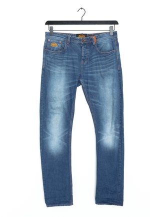 Jean en denim bleu avec les cuisses et les genoux avant décolorés suspendu sur un cintre noir sur fond blanc.