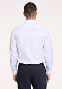 Camisa blanca de manga larga con cuello abotonado, que presenta una textura suave y un brillo sutil, combinada con pantalones oscuros de color azul marino.