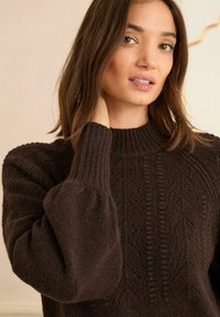 Brauner, gestrickter Pullover mit hohem Rippkragen, strukturiertem Muster und voluminösen Ärmeln mit Rippbündchen. Weicher, warmer Stoff.