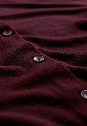 Burgunderfarbener Strickcardigan mit glatter Textur, auffälligen Knopfdetails und dezenten Drapierungen. Die Knöpfe sind dunkel, rund und glänzend.
