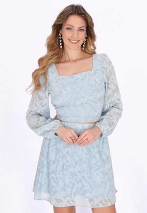 Žena v světle modrých květovaných krajkových šatech s dlouhými nafouknutými rukávy a hranatým výstřihem, usmívající se s rukama sepnutýma před sebou.