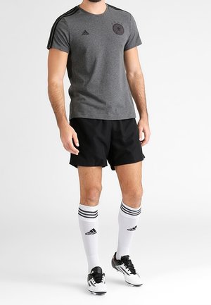Camiseta deportiva gris de manga corta, con logo negro, combinada con pantalones cortos negros y calcetas blancas hasta la rodilla con rayas negras, que presenta tacos.