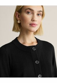 Musta pehmeästä kankaasta valmistettu neuletakki, jossa pyöreä pääntie, neljä mustaa nappia ja yksinkertainen, koristeeton muotoilu.