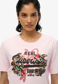 Camiseta de algodón rosa con un diseño adornado con lentejuelas de peces koi y texto en negro, rojo y verde. Manga corta y cuello redondo.