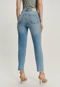 Mujer vestida con jeans de tiro alto azul claro y sandalias de tacón alto beige con tiras, de pie frente a un fondo liso.