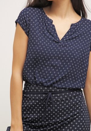 Femme portant un haut à manches courtes bleu marine à motifs et une jupe assortie avec de petites formes géométriques blanches, nouée à la taille avec un cordon.