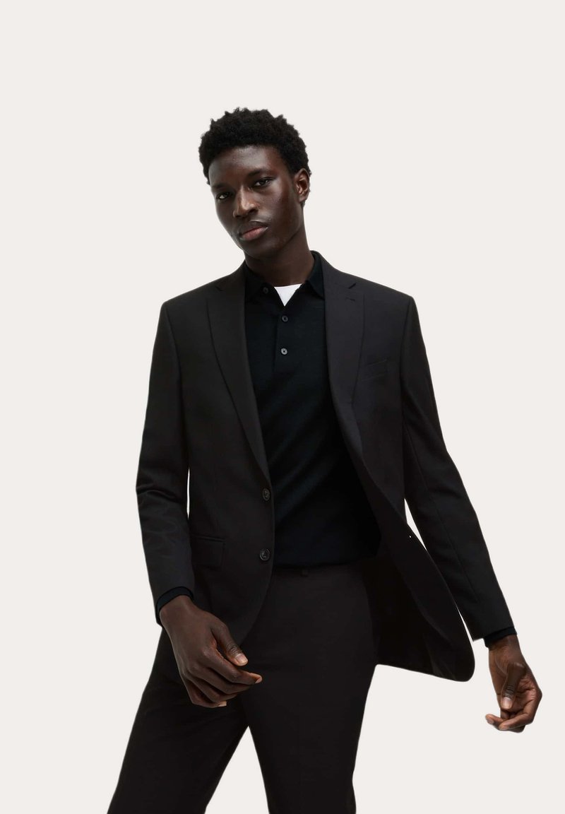 Czarny garnitur o dopasowanym kroju z gładką teksturą, z dwoma guzikami, klapami w kształcie notcha i dopasowanymi spodniami. Noszony na czarnej koszulce polo.