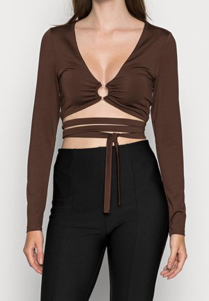 Kvinde iført en brun langærmet crop top med en central ringdetalje og omslagstropper, kombineret med højtaljede sorte bukser.