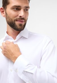 Witte button-down shirt met lange mouwen, een puntige kraag en een gladde textuur. Weergegeven met een hand die de kraag vastmaakt.