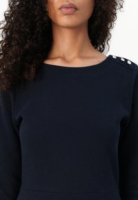 Tmavě modré texturované šaty s lodičkovým výstřihem, zdobené stříbrnými knoflíky na rameni a dlouhými rukávy.