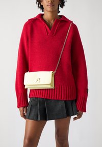 Pull en tricot rouge avec un large col et un ourlet côtelé, associé à une jupe plissée à carreaux noire. Un petit sac bandoulière jaune avec une chaîne.