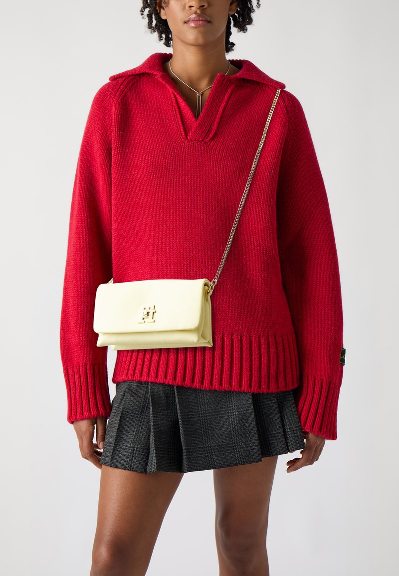 Pull en tricot rouge avec un large col et un ourlet côtelé, associé à une jupe plissée à carreaux noire. Un petit sac bandoulière jaune avec une chaîne.