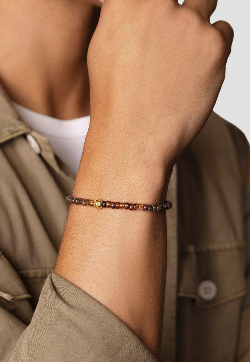 Beaded bracelet featuring multicolored glass beads in amber and brown tones, with a small gold accent bead. Worn on a wrist.