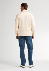 Chaqueta sudadera beige con cuello tipo camisa, hombros caídos y un ajuste ligeramente holgado, combinada con jeans de mezclilla azules y zapatillas blancas.