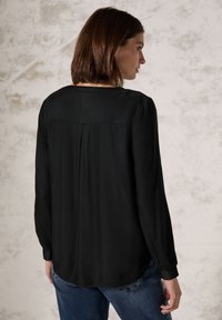 Blusa nera a maniche lunghe con una texture liscia, caratterizzata da una piega sul retro e un orlo arrotondato, abbinata a jeans blu.