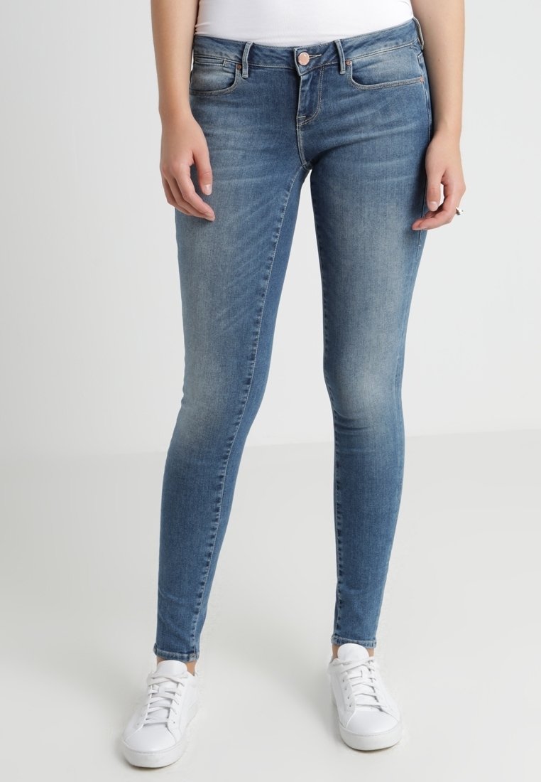 Kvinna som bär blåa slim fit-jeans och vita sneakers, står mot en neutral ljus bakgrund, med avslappnade händer vid sidorna.