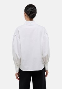 Chemise en coton blanc avec de longues manches bouffantes et des poignets à boutons. Présente un dos avec yoke et un col classique. Tissu lisse et structuré.