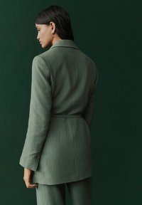 Hellgrüner, strukturierter Blazer mit langen Ärmeln und gebundenem Taillengürtel, mit spitzem Kragen und Knopf am Manschette, vor einem dunkelgrünen Hintergrund.