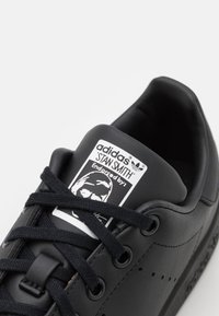 Sort læder adidas Stan Smith sneakers med flade snørebånd, perforerede detaljer og et mærke på tungen med et portræt grafik.