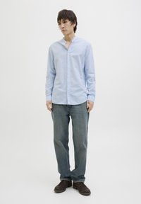 Camisa de botones azul claro con cuello mao, de manga larga, combinada con vaqueros de mezclilla azul medio y zapatos marrón oscuro.