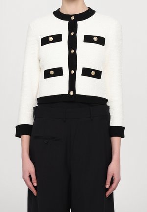 Weisse kurze Jacke mit schwarzem Besatz und goldenen Knöpfen, getragen mit schwarzen Hosen mit hoher Taille.