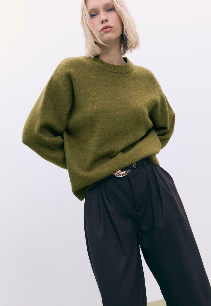 Oliwkowy, oversize sweter z długimi rękawami, z prążkowanym dekoltem. Połączony z czarnymi spodniami o wysokim stanie z plisami i metalowym paskiem.
