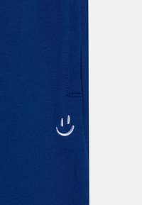 Námořnické modré bavlněné kalhoty s bílou vyšívanou usměvavou tváří na levé kapse. Měkká textura a neformální design.