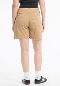 Shorts in cotone khaki con orlo sfrangiato, due tasche posteriori e un piccolo logo vicino alla cintura. Indossati con sneakers nere e calzini bianchi.