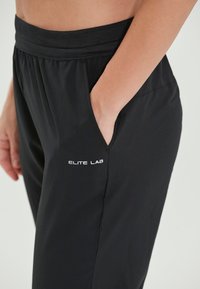 Sorte træningsbukser lavet af letvægtsstof med elastisk talje, udstyret med sidelommer og et hvidt "ELITE LAB" logo.