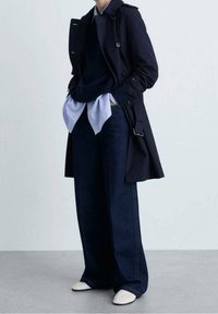 Osoba oblečená v vrstveném námořnickém trenčkotě, tmavých širokých kalhotách, světle modré košili, námořnickém svetru a bílých botách, stojící s rukama v kapsách.