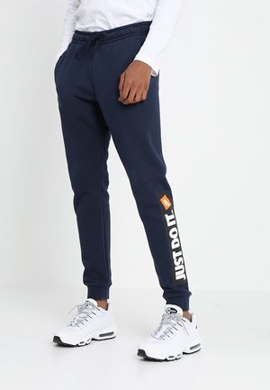 Pantalons de jogging bleu marine avec une taille élastique, poches latérales, et le texte "JUST DO IT." sur la jambe gauche. Associés à des baskets blanches.