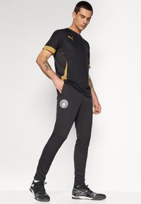 Sort sportstrøje med guld accenter og logo; parret med sorte tapered joggers med et cirkulært logo på venstre lår og sorte sneakers.