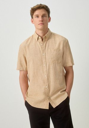 Fiatal férfi, bézs színű rövid ujjú, mellzsebes gombos ingben és fekete nadrágban, kezeit a zsebében tartva, egyszerű háttér előtt áll.