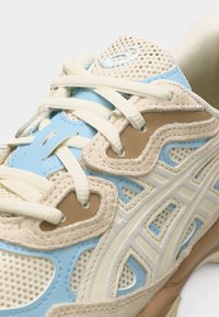 Gros plan d'une basket beige et bleu clair avec des panneaux en mesh, des lacets crème et des détails cousus.
