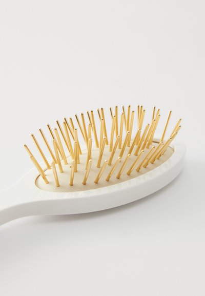 Οβάλ βούρτσα μαλλιών με λευκή λαβή, που διαθέτει χρυσές μεταλλικές τρίχες σε ανάγλυφη κρεμ βάση. Απλός και εργονομικός σχεδιασμός.