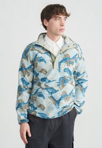 Νέος άνδρας που φοράει fleece πουλόβερ με μπλε, μπεζ και λευκό μοτίβο πάνω από λευκό πουκάμισο με κουμπιά και σκούρο παντελόνι, στέκεται μπροστά σε απλό φόντο.