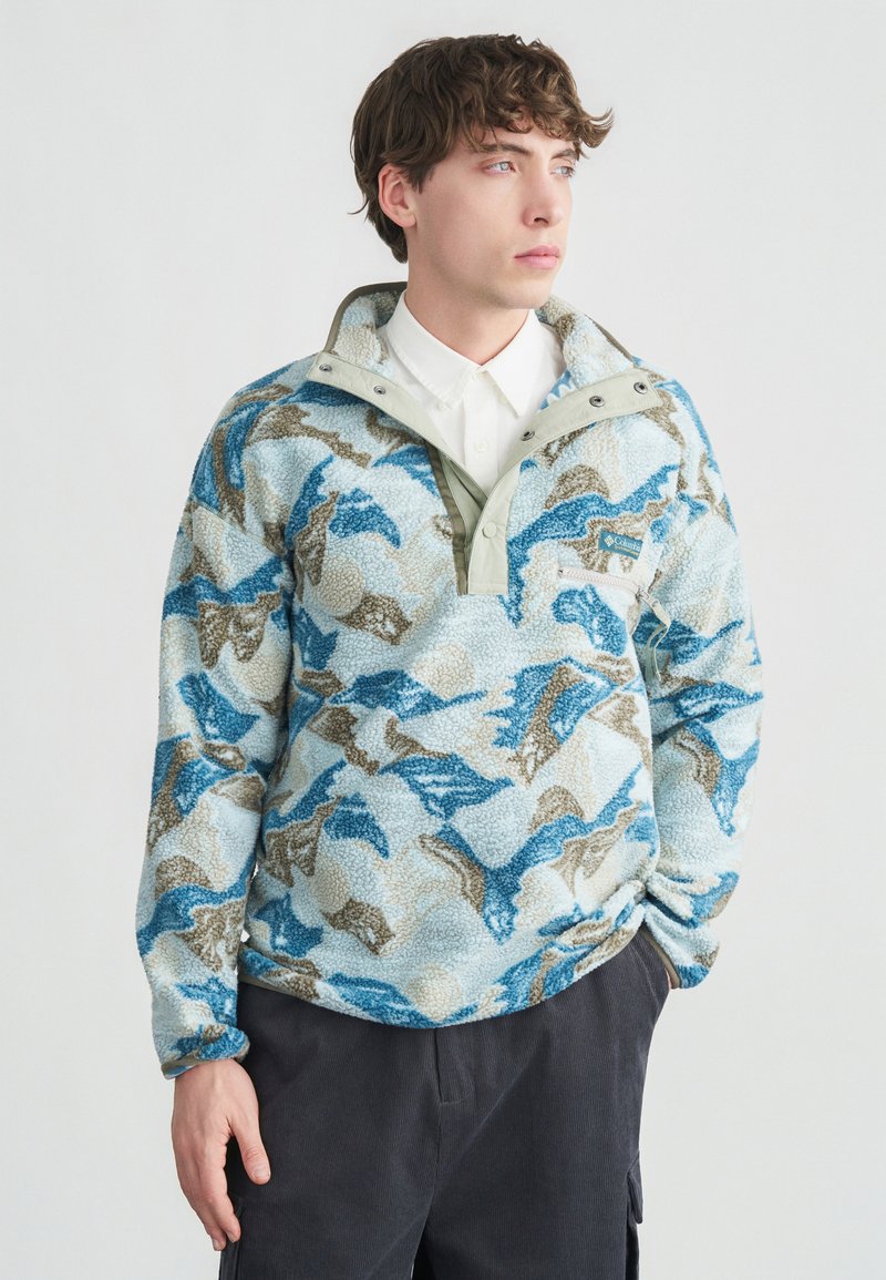 Νέος άνδρας που φοράει fleece πουλόβερ με μπλε, μπεζ και λευκό μοτίβο πάνω από λευκό πουκάμισο με κουμπιά και σκούρο παντελόνι, στέκεται μπροστά σε απλό φόντο.