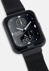 Reloj inteligente negro con una cara cuadrada, acentos plateados, pantallas analógicas y digitales, y una correa de malla metálica texturizada. Incluye información de fecha y del tiempo.