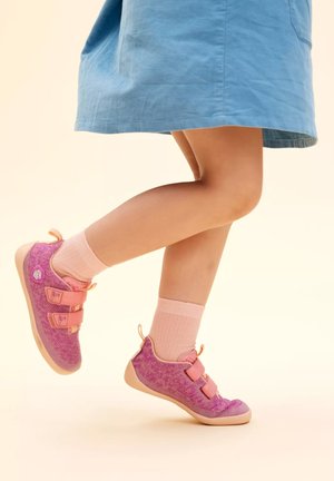 Paarse en roze schoenen met een structuurgebreide bovenkant, twee klittenbandsluitingen en een lichte zool; gecombineerd met roze enkelsokken en een blauwe jurk.
