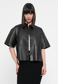 Svart lær skjortejakke med korte ermer, knappestolpe og klassisk krage, med en glatt tekstur og strukturert design.