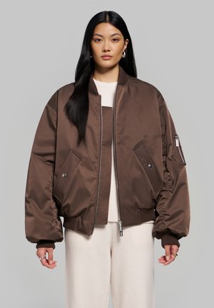 Femme aux longs cheveux noirs portant une veste bomber marron oversize, un pull crème et un pantalon crème, debout devant un fond gris uni.