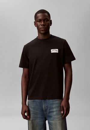 Férfi fekete Calvin Klein pólóban és kifakult kék farmerben, egyszínű szürke háttér előtt áll.