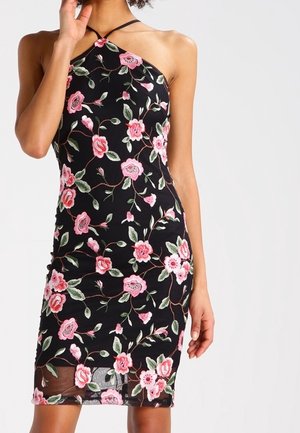 Sort halterneck-kjole med lyserød og grøn blomstermotiv broderi, tætsiddende silhuet, båret af en person med den ene hånd, der rører ved ansigtet.