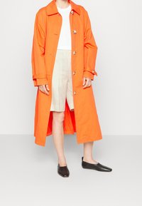 Oranžový bavlněný trenčkot s klasickým límcem, předním zapínáním na knoflíky a ohrnutými rukávy. Nositý přes bílý top a béžové šortky, s černými balerínami.