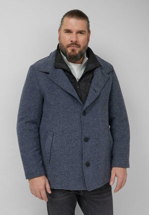 Uomo barbuto con capelli legati indietro, indossa un cappotto di lana grigio sopra una giacca nera con cerniera e una camicia bianca, in piedi davanti a uno sfondo neutro.